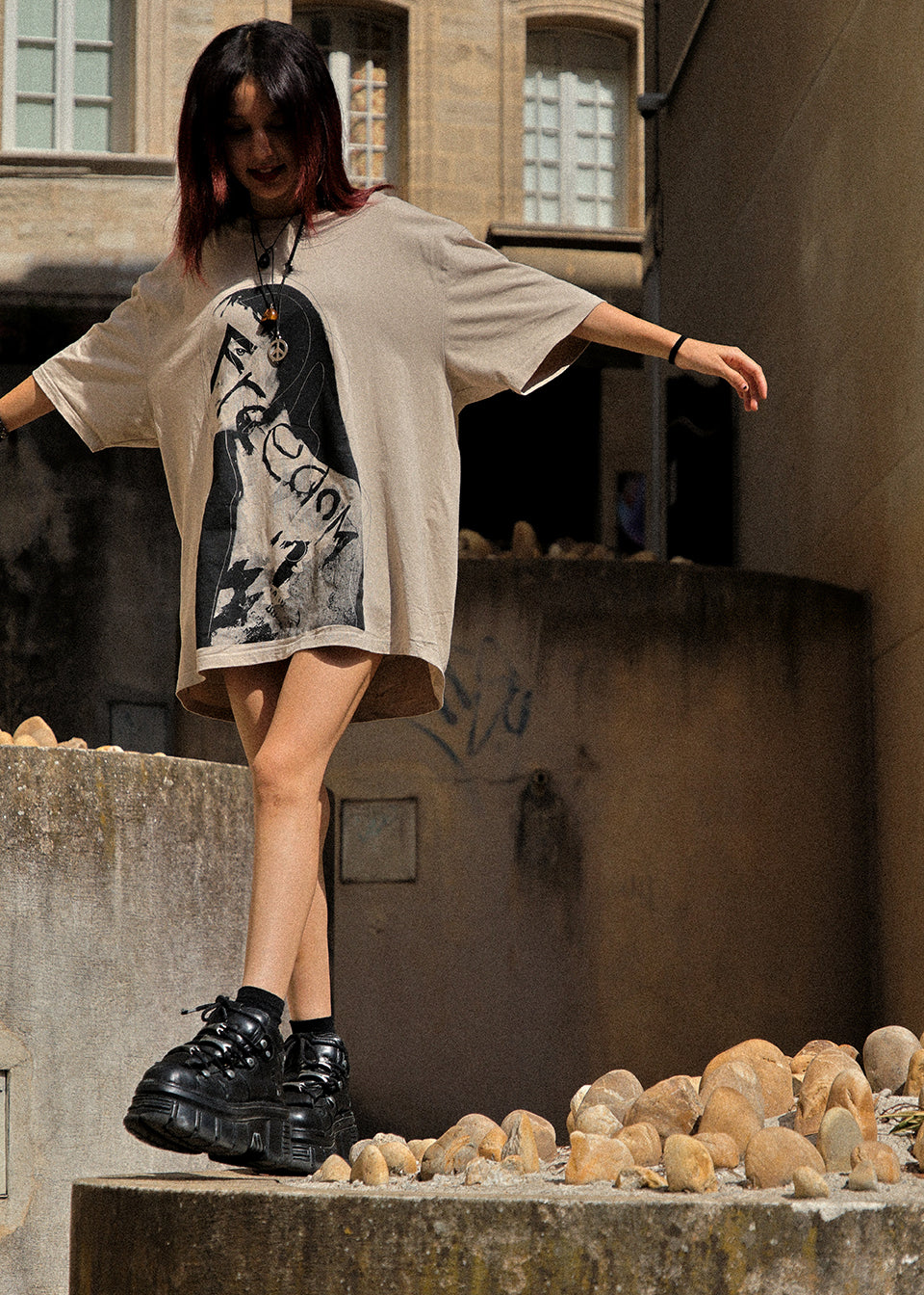 A model wearing an oversized beige graphic t-shirt dress with a black and white print, paired with chunky black platform boots, posing on a stone ledge in an urban setting.
