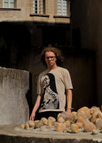 A person with curly hair and glasses stands outdoors in front of a stone wall, wearing a beige graphic T-shirt with a black-and-white portrait print and black shorts. Rocks are piled in the foreground.