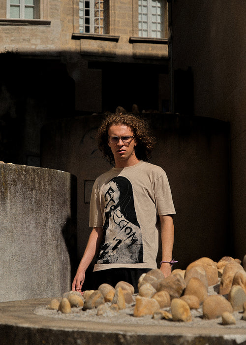 A person with curly hair and glasses stands outdoors in front of a stone wall, wearing a beige graphic T-shirt with a black-and-white portrait print and black shorts. Rocks are piled in the foreground.