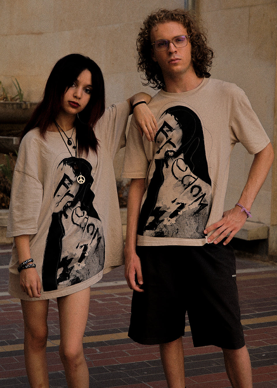 Two models wearing oversized beige graphic t-shirts featuring a black and white portrait of a woman with the text 'Fiction' and 'Fiction 1', standing outdoors against a stone wall.