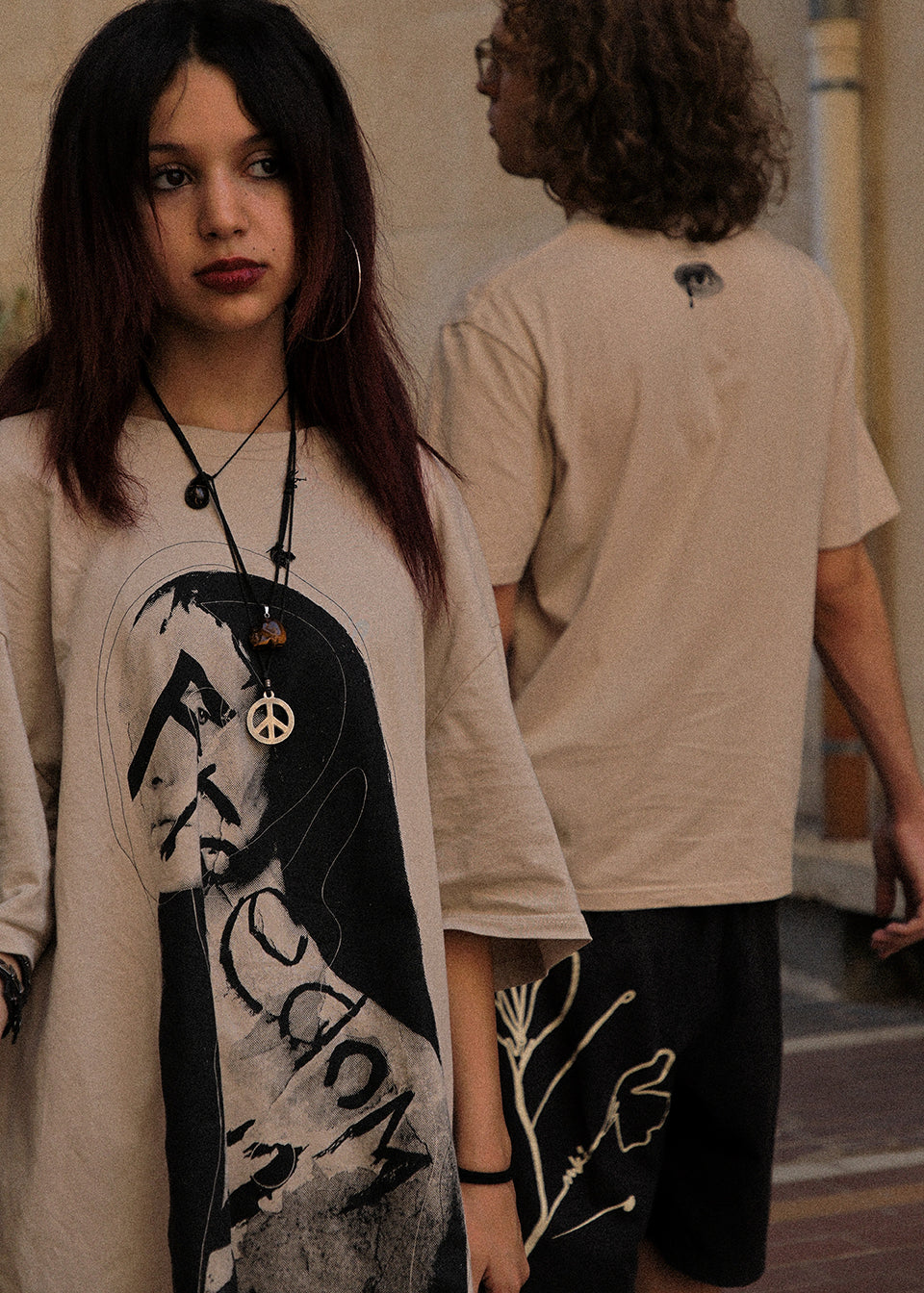 A person wearing a beige graphic T-shirt with a large black-and-white portrait print and a peace sign pendant necklace, standing in an outdoor setting. Another person in a beige T-shirt and dark shorts is blurred in the background.