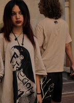A person wearing a beige graphic T-shirt with a large black-and-white portrait print and a peace sign pendant necklace, standing in an outdoor setting. Another person in a beige T-shirt and dark shorts is blurred in the background.