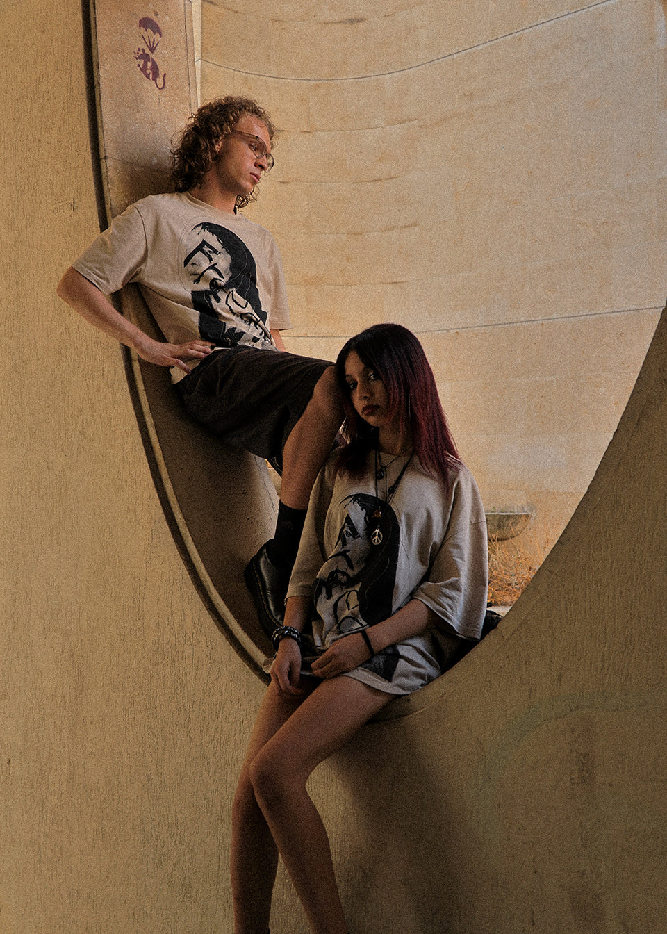Two models posing in a concrete bowl structure, wearing matching graphic t-shirts with a black-and-white portrait print and dark shorts.