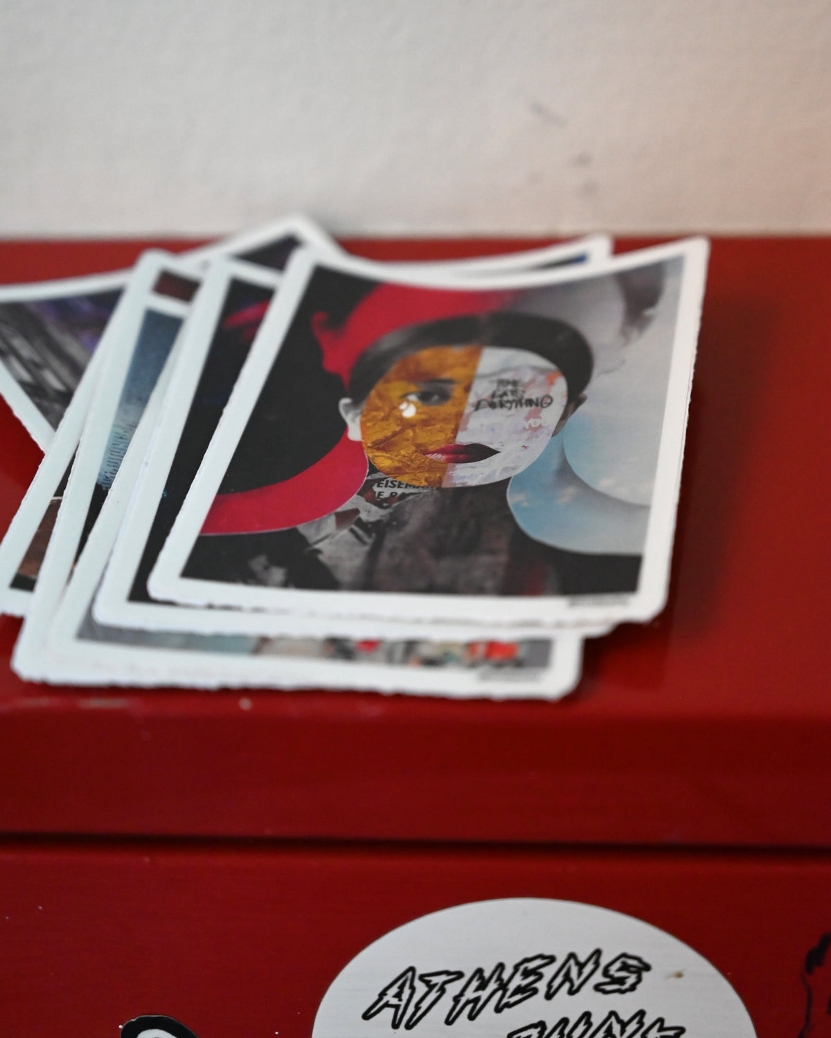 A stack of printed art cards or postcards with a collage portrait on the top card, resting on a red surface against a light wall.
