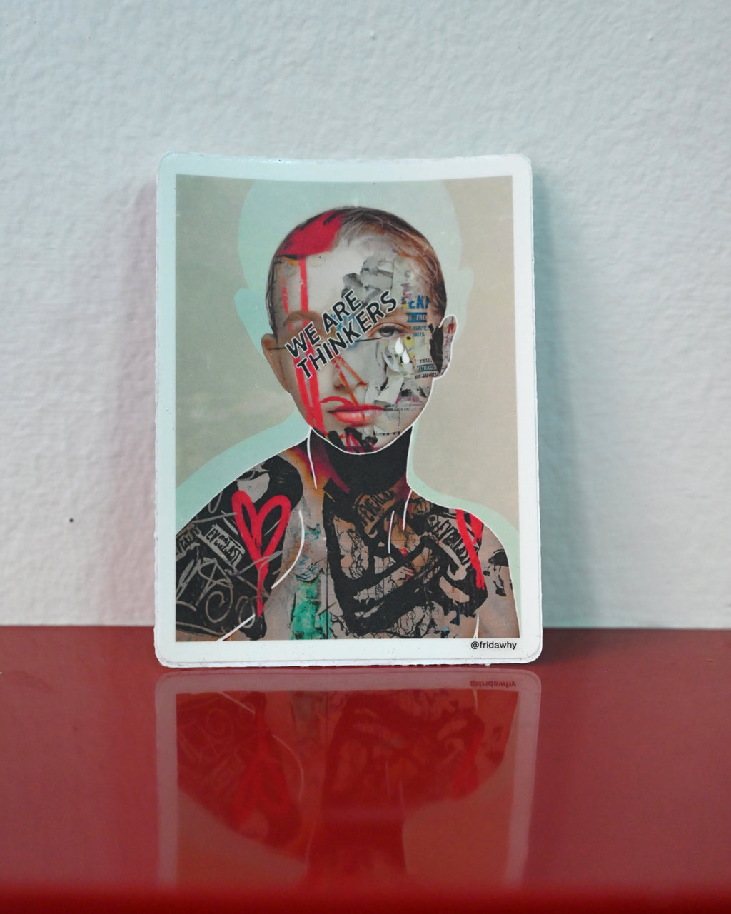 A rectangular art print or sticker with rounded corners featuring a stylized portrait of a tattooed person with red paint and the text 'WE ARE THINKERS', displayed upright on a glossy red surface against a light gray wall.