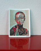 A rectangular art print or sticker with rounded corners featuring a stylized portrait of a tattooed person with red paint and the text 'WE ARE THINKERS', displayed upright on a glossy red surface against a light gray wall.