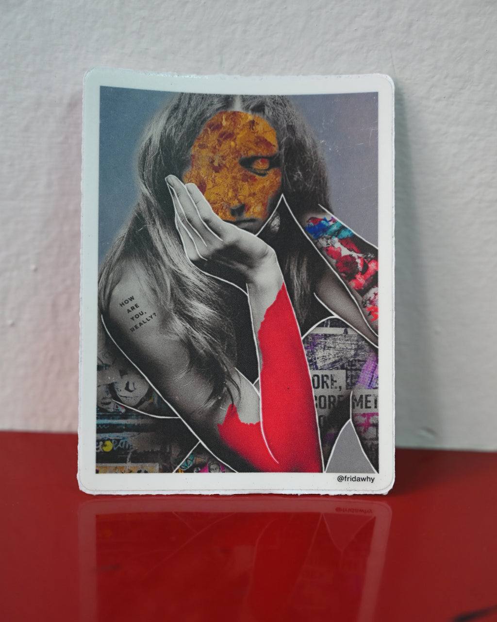 A rectangular art print with a white border featuring a collage portrait of a woman with a textured orange-yellow face, black-and-white hair, and a red garment, leaning against a light wall on a glossy red surface.