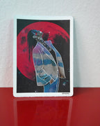 A rectangular art print with a white border stands upright on a glossy red surface against a white wall. The artwork shows a stylized figure in a light blue blazer against a large red moon on a dark background, with the words 'You Are Not Alone' across the neck area.