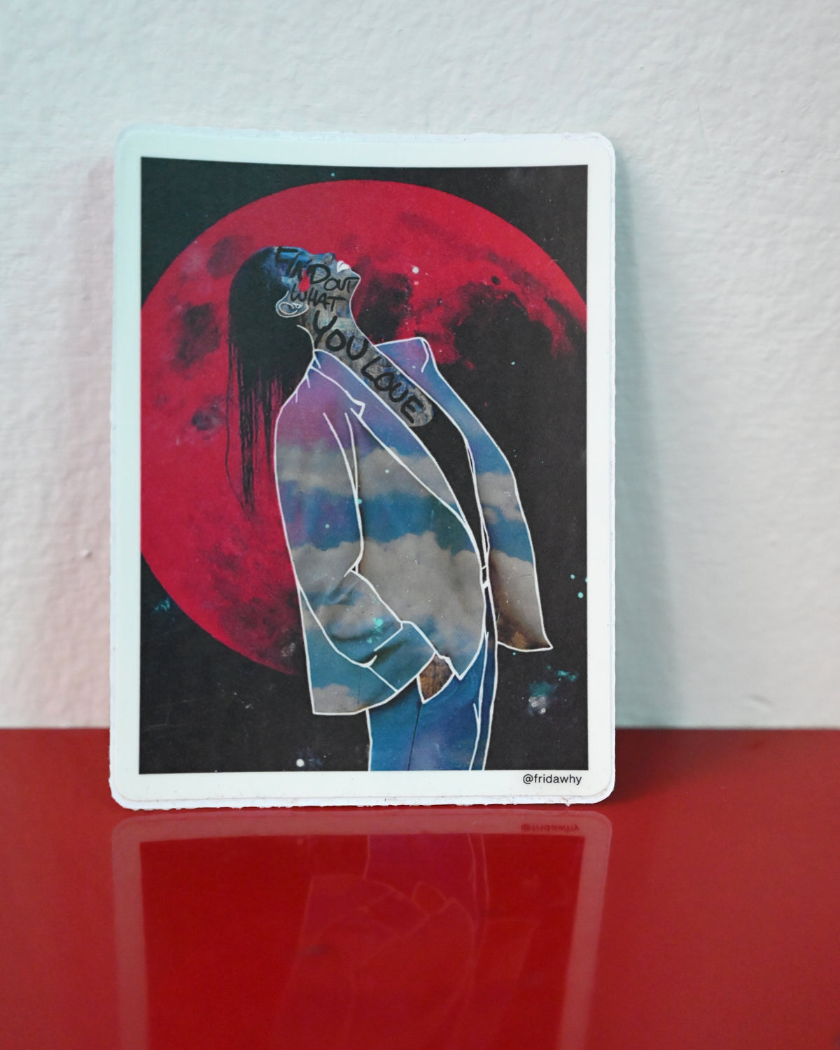 A rectangular art print with a white border stands upright on a glossy red surface against a white wall. The artwork shows a stylized figure in a light blue blazer against a large red moon on a dark background, with the words 'You Are Not Alone' across the neck area.