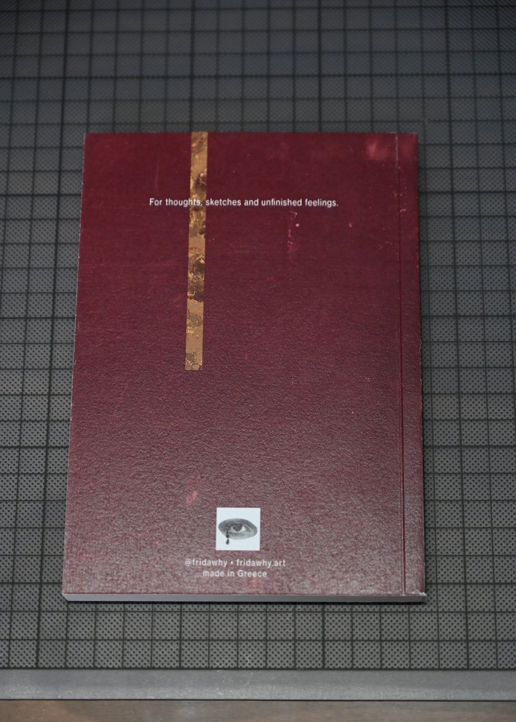 A square dark red textured book or journal with a vertical gold stripe down the center, placed on a dark grid cutting mat. White text near the top reads 'For thoughts, sketches and unfinished feelings.