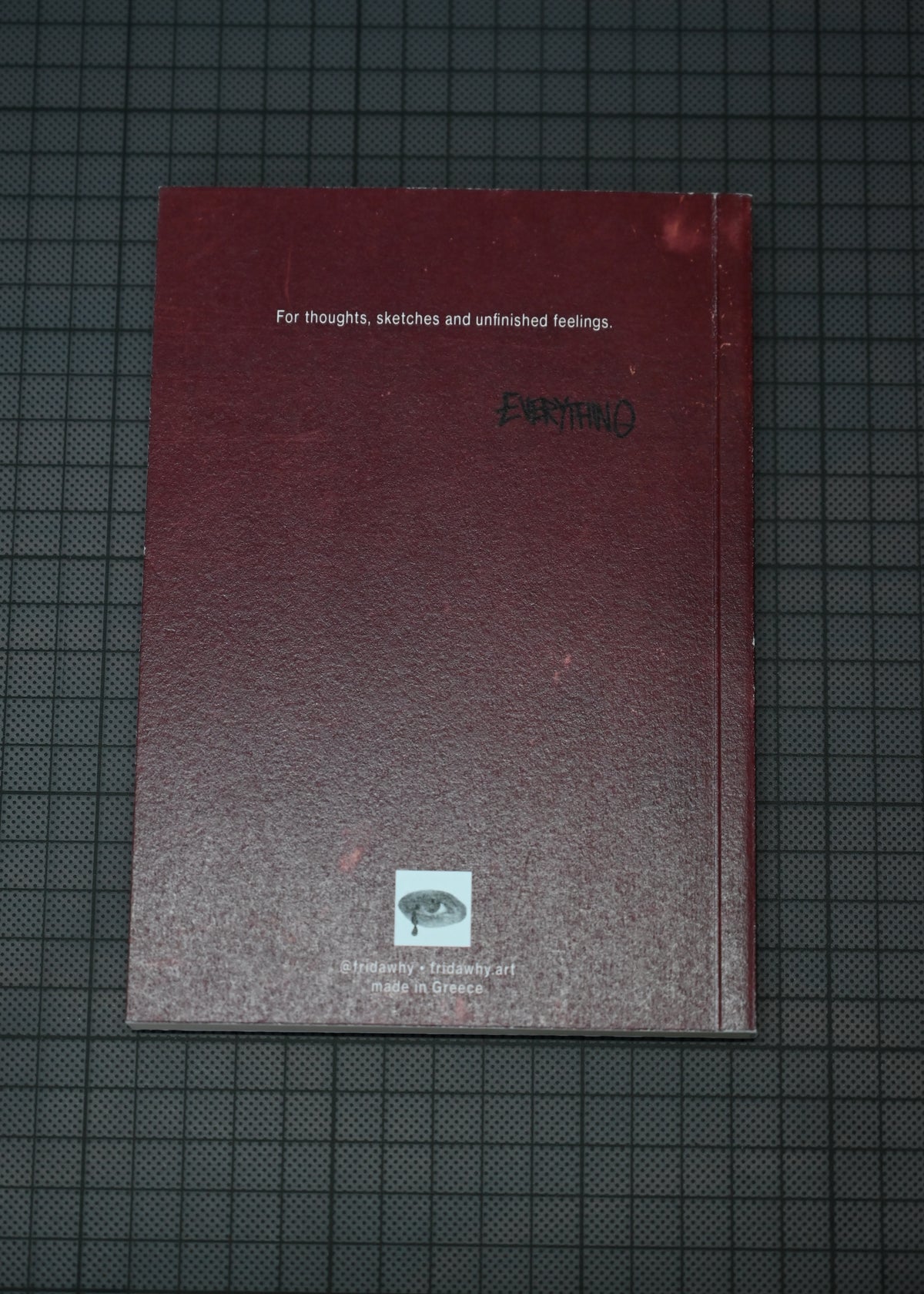 A dark red sketchbook or notebook with white text reading 'For thoughts, sketches and unfinished feelings.' and a small eye logo near the bottom, placed on a dark gridded surface.