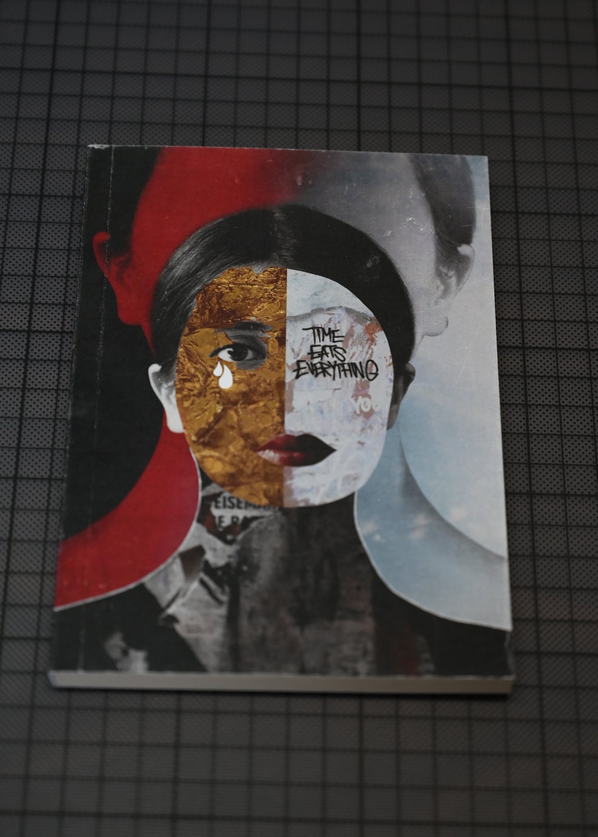 A square art print or book cover featuring a stylized portrait split into black, white, red, and textured gold sections, placed on a dark gridded surface.