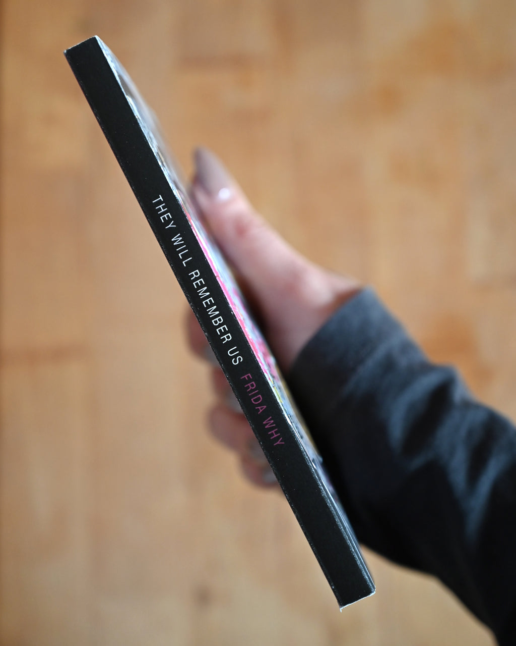 Hand holding a book titled 'They Will Remember Us Frida' on a wooden surface