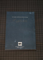 A dark slate blue sketchbook lying flat on a dark grid-patterned surface. The cover features white text at the top, faint handwritten text in the middle, a small eye logo near the bottom, and social media details underneath.