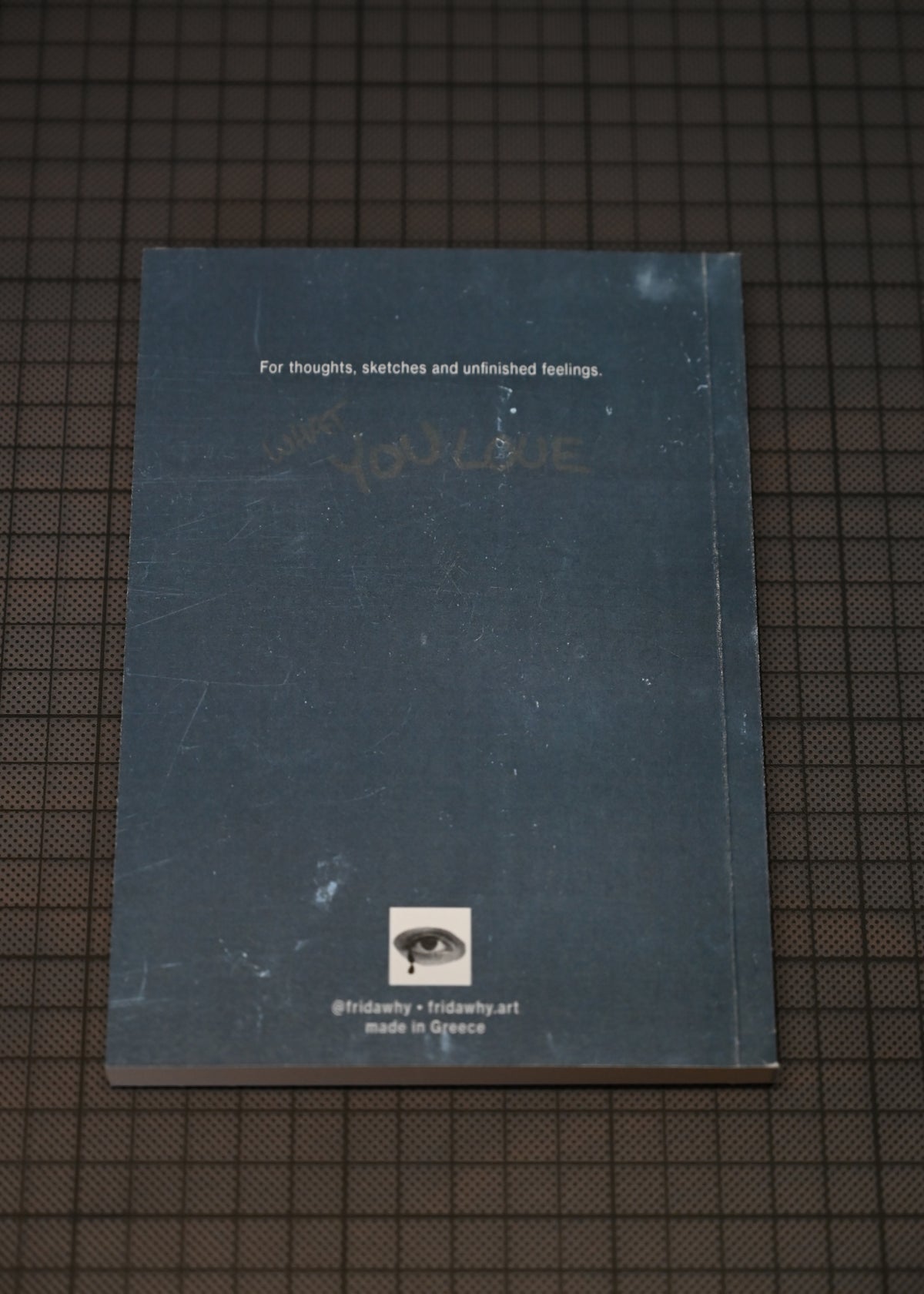A dark slate blue sketchbook lying flat on a dark grid-patterned surface. The cover features white text at the top, faint handwritten text in the middle, a small eye logo near the bottom, and social media details underneath.
