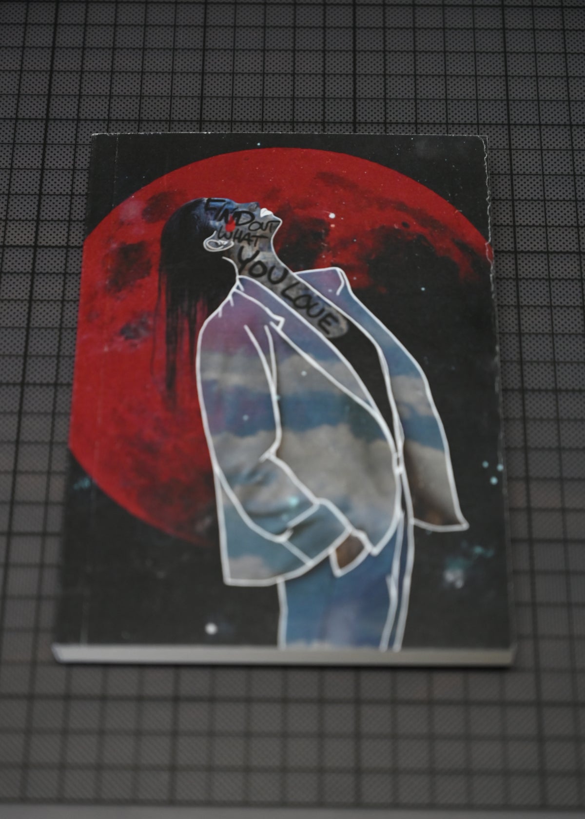A rectangular art print or book cover featuring a stylized figure in a white coat in front of a large red moon on a dark starry background, photographed on a gray grid surface.