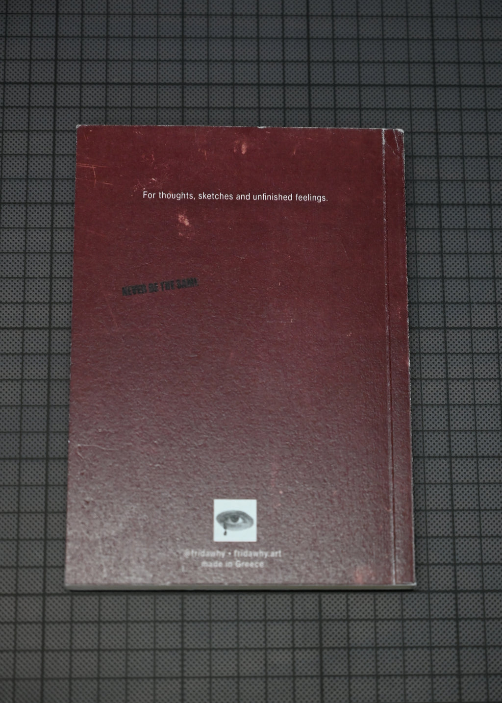 A dark red textured notebook or journal with white text and a small eye logo near the bottom, placed on a dark grid-patterned surface.