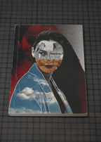 A rectangular printed artwork or book-like item lying on a dark grid surface, featuring a stylized portrait collage with a woman's face layered over red, black, blue sky, and textured earth tones.