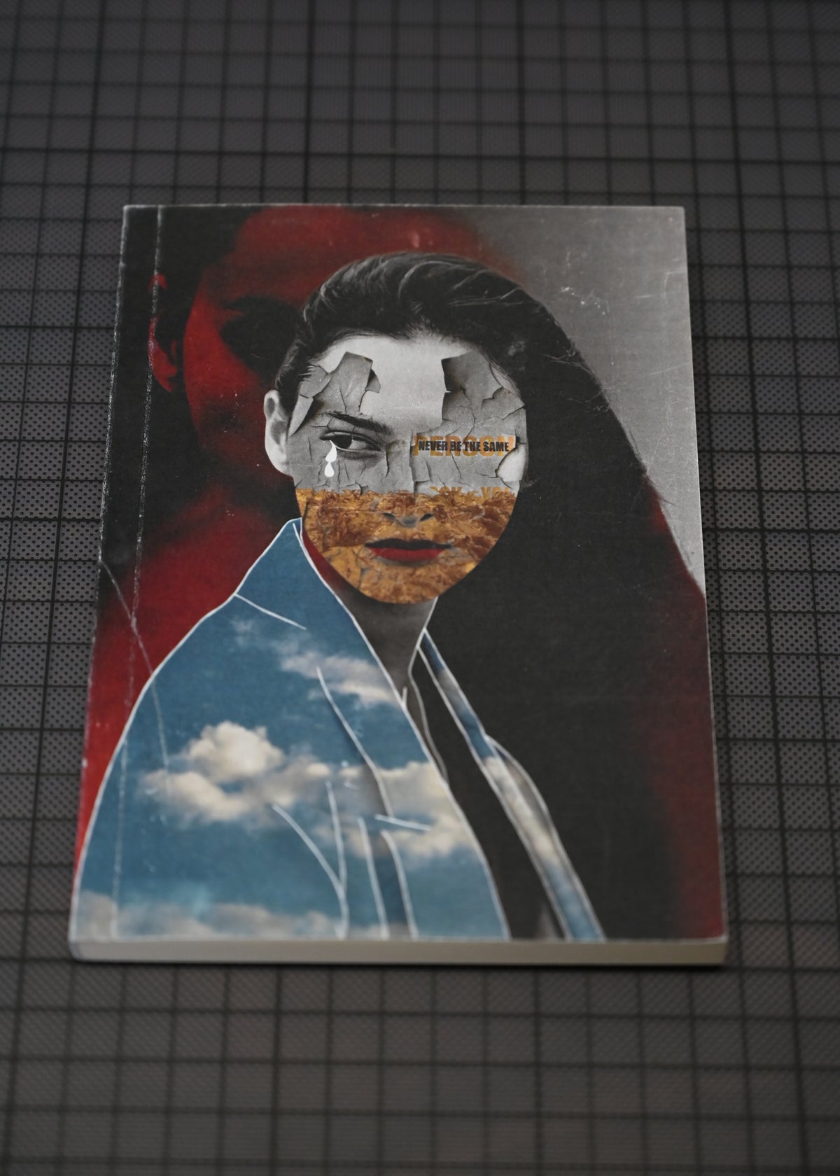 A rectangular printed artwork or book-like item lying on a dark grid surface, featuring a stylized portrait collage with a woman's face layered over red, black, blue sky, and textured earth tones.