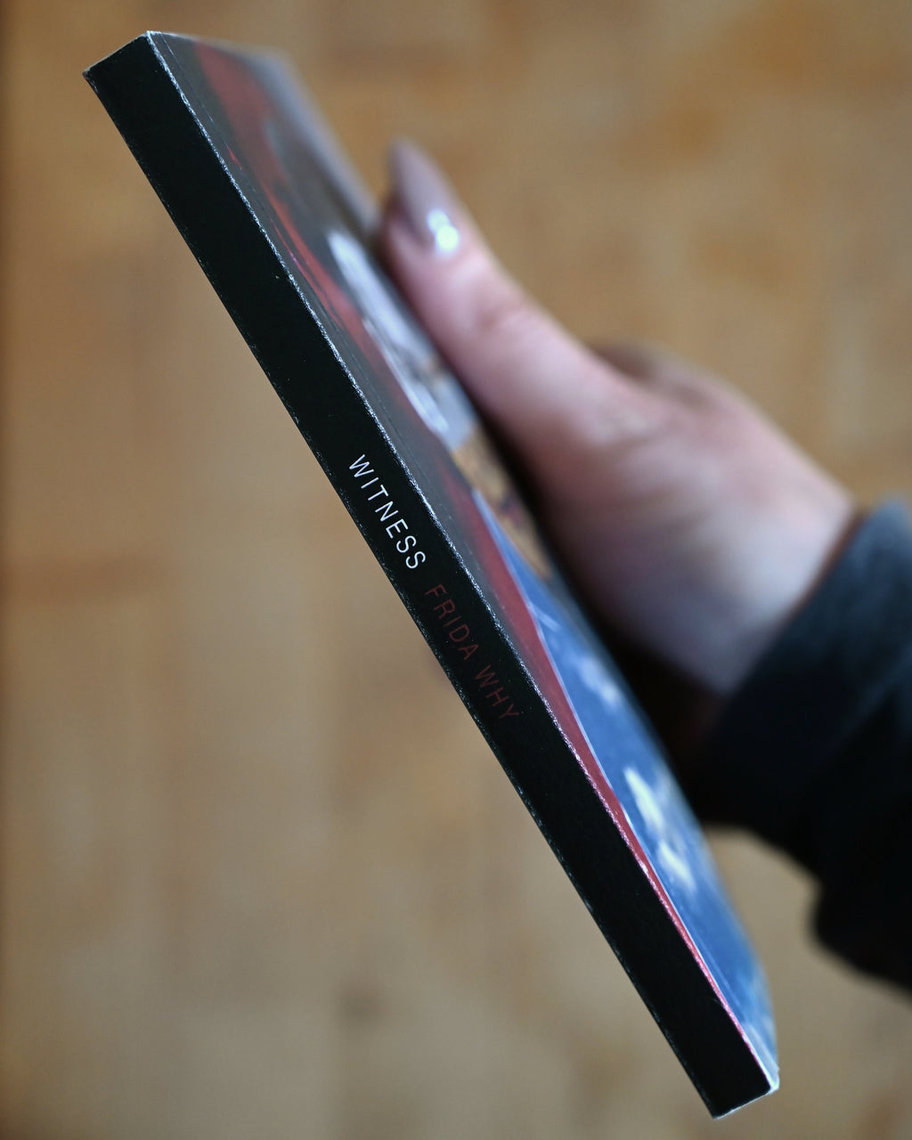 Hand holding a black and red product with 'WITNESS' text on a wooden surface