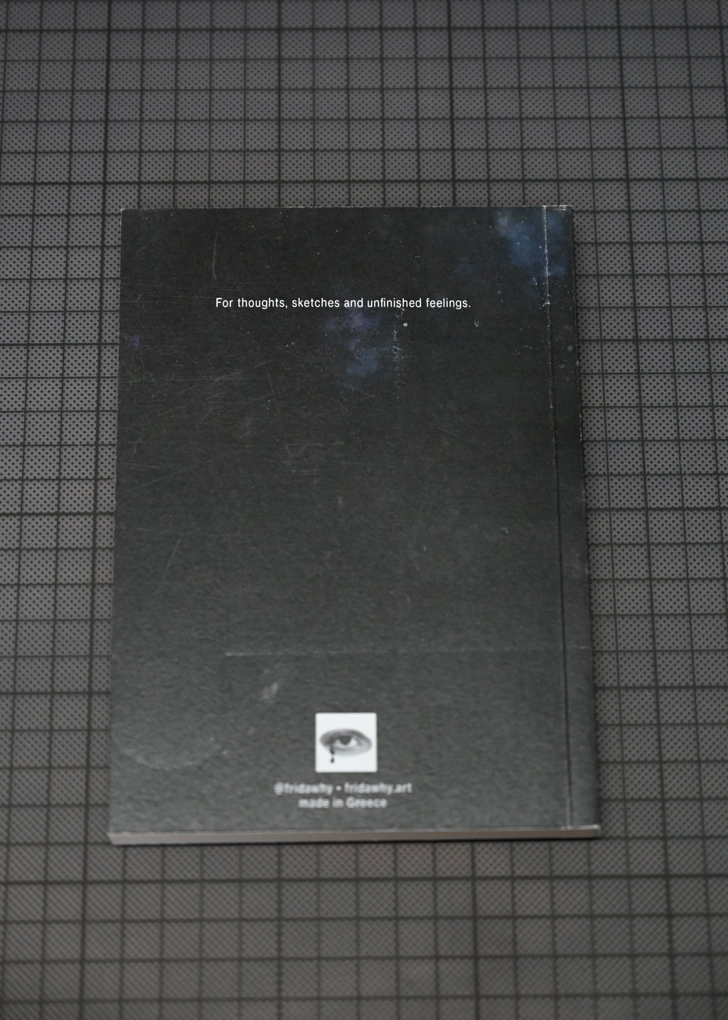 A black sketchbook or notebook with white text and a small logo near the bottom, placed on a dark grid-patterned surface.