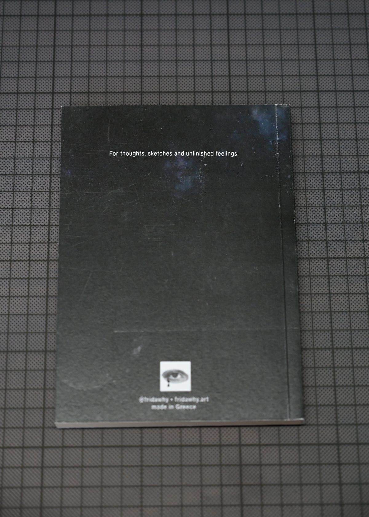 A black sketchbook or notebook with white text and a small logo near the bottom, placed on a dark grid-patterned surface.