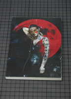 A hardcover notebook or journal lying flat on a black grid cutting mat. The cover features a dark, artistic illustration of a tattooed woman with red eyes in front of a large red moon and a starry galaxy background.