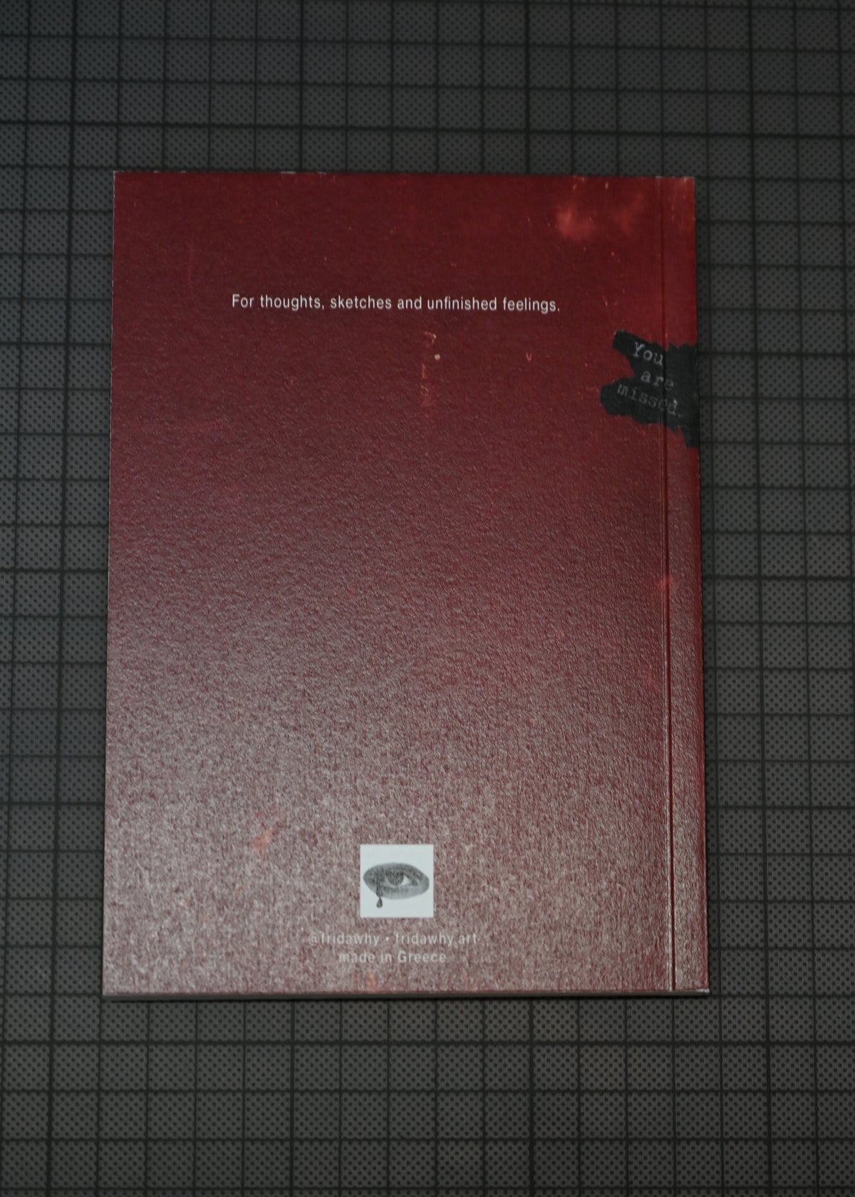 A textured red sketchbook or notebook with white text reading 'For thoughts, sketches and unfinished feelings.' lies on a dark grid-patterned surface.