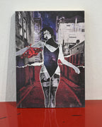 A rectangular art print or poster featuring a stylized collage of a woman in a black bodysuit standing in a red-lit alley, with torn-paper effects and a red abstract shape on the left, displayed against a light wall on a red surface.