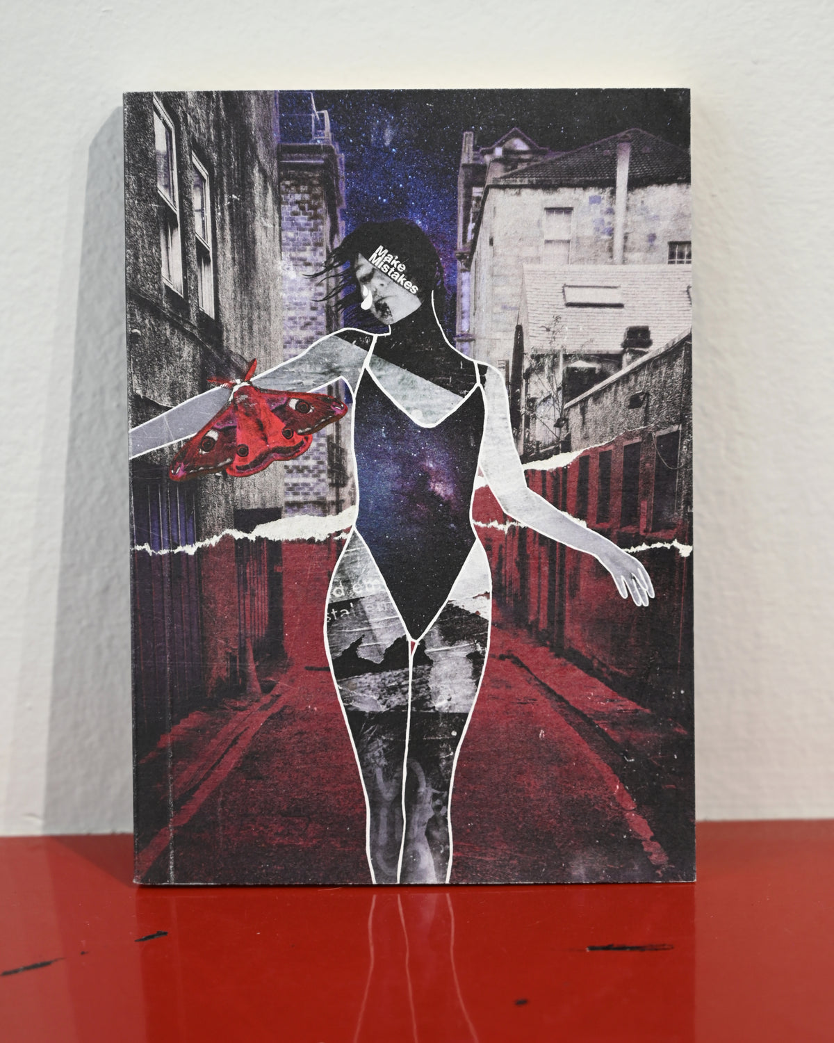A rectangular art print or poster featuring a stylized collage of a woman in a black bodysuit standing in a red-lit alley, with torn-paper effects and a red abstract shape on the left, displayed against a light wall on a red surface.