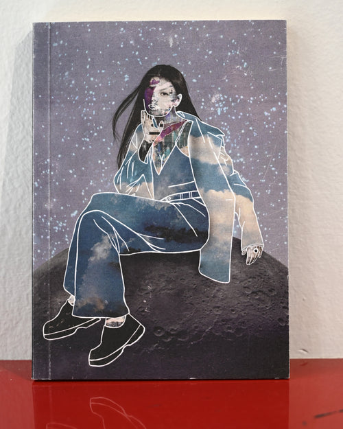 A hardcover notebook or journal standing upright on a red reflective surface against a white wall. The cover features an illustrated portrait of a seated woman with long dark hair, wearing a light blue jacket and pants, set against a starry purple sky and a dark moon-like surface.