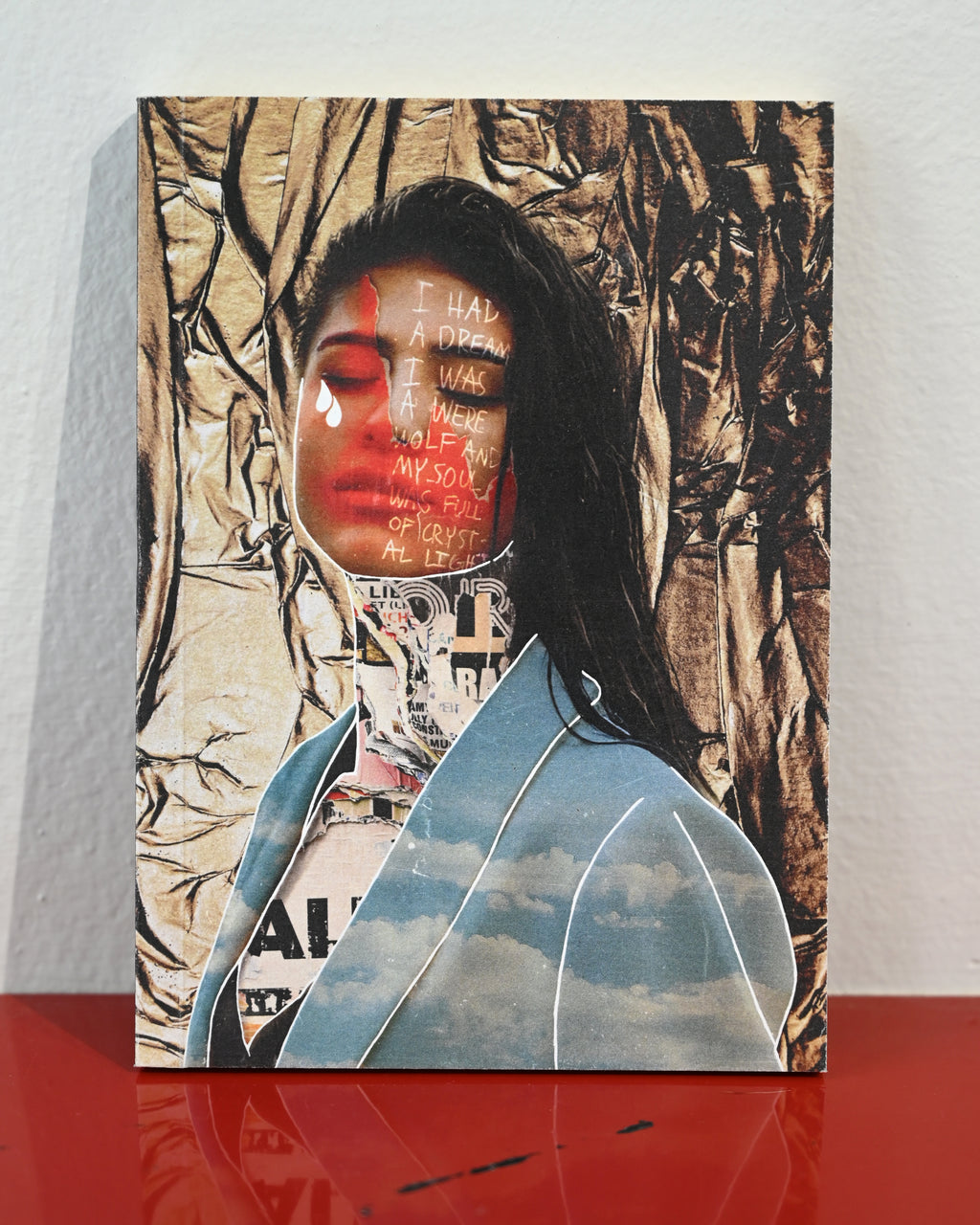 A vertical mixed-media collage art print depicting a woman with dark hair, red lips, and text overlays, styled with a light blue jacket and a sky pattern, displayed on a red reflective surface against a white wall.
