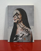 A square art print or canvas featuring an abstract portrait of a woman with long black hair, set against a gray textured background with black graphic lines and text, displayed on a red reflective surface against a white wall.