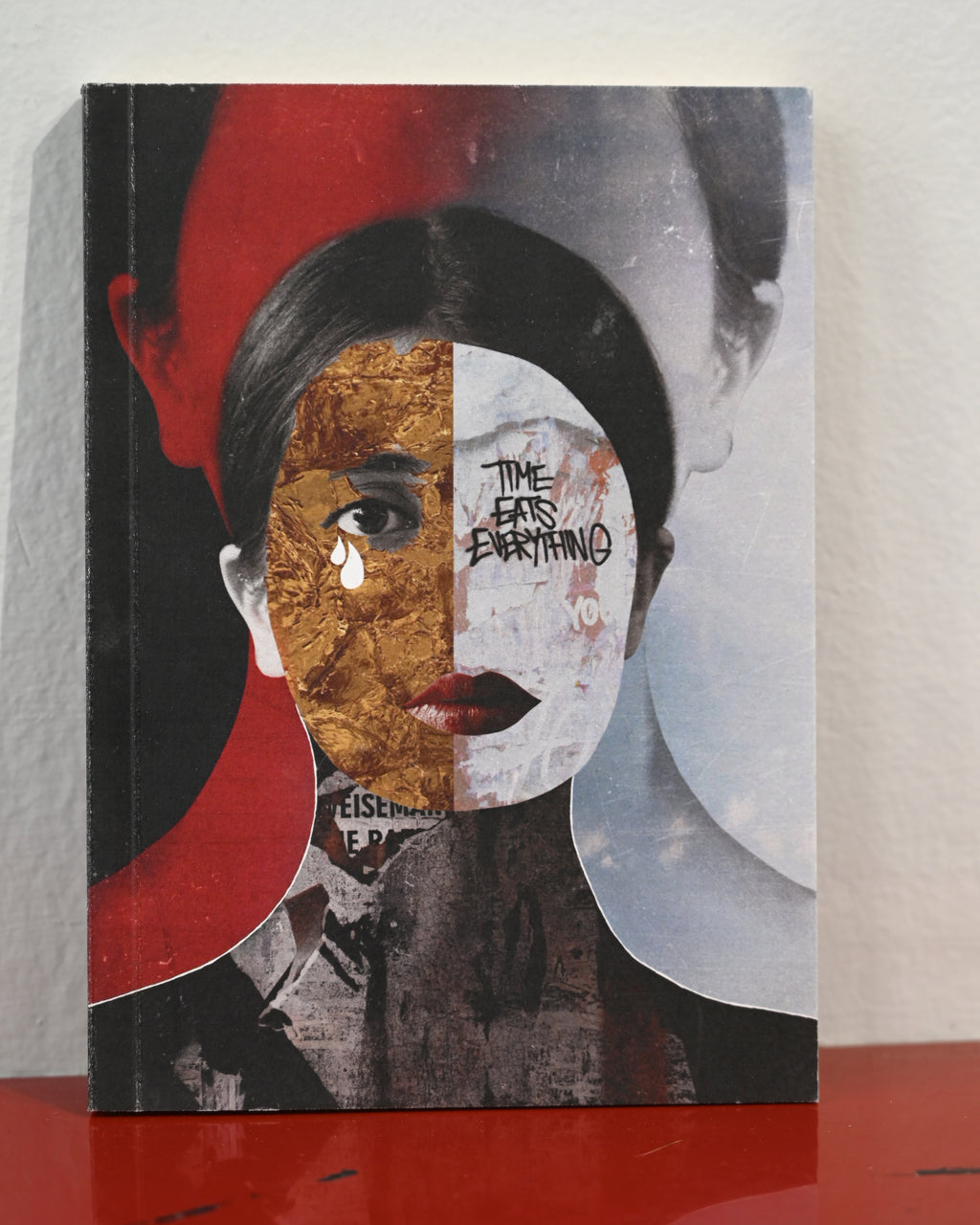 A square art print or book cover featuring a stylized portrait collage with a woman's face split into black, red, gold, white, and gray sections, standing on a red surface against a light wall.