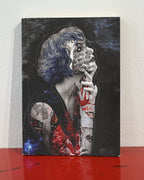 A hardcover art journal standing upright on a glossy red surface against a white wall. The cover features a dark collage portrait of a woman with blue hair, tattoos, and handwritten text, with a small blue galaxy graphic in the lower left corner.