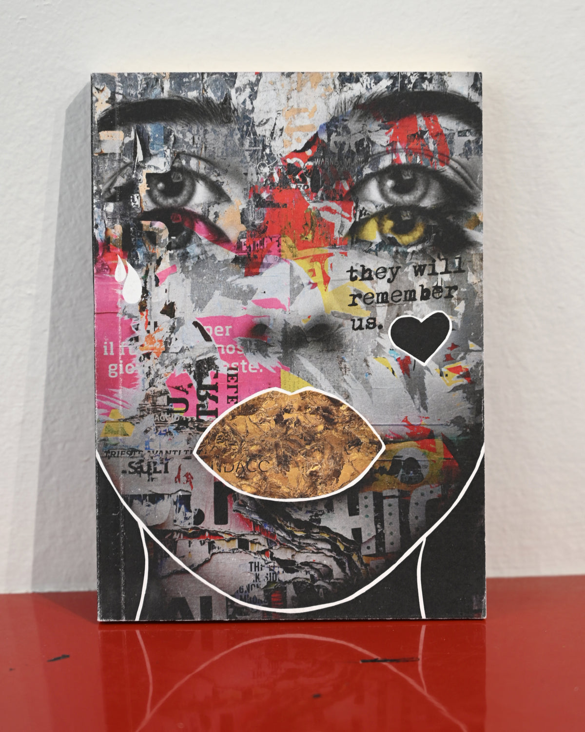 A mixed-media collage art print featuring a stylized face with cutout eyes and lips, displayed on a red reflective surface against a white wall.