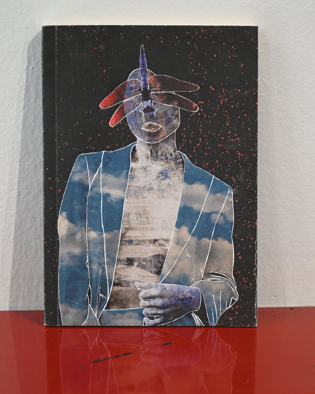 A rectangular mixed-media art print or canvas featuring a surreal figure with a blue and white cloud-patterned jacket, a metallic blue face, and red hat accents on a black speckled background, displayed standing on a glossy red surface against a white wall.