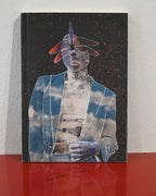 A rectangular mixed-media art print or canvas featuring a surreal figure with a blue and white cloud-patterned jacket, a metallic blue face, and red hat accents on a black speckled background, displayed standing on a glossy red surface against a white wall.