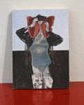 A square art print or book cover featuring a surreal collage of a figure in black clothing with a large red bow or headpiece, set against a pale blue patterned background and a dark lower section. The print is photographed standing on a glossy red surface against a light wall.
