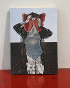 A square art print or book cover featuring a surreal collage of a figure in black clothing with a large red bow or headpiece, set against a pale blue patterned background and a dark lower section. The print is photographed standing on a glossy red surface against a light wall.