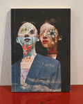 A hardcover art book or journal standing upright on a red surface against a light beige wall. The cover features a surreal collage portrait of two women with mixed media elements including graffiti, a red fox, and a flower motif, with a blue sky and clouds printed over the lower portion.