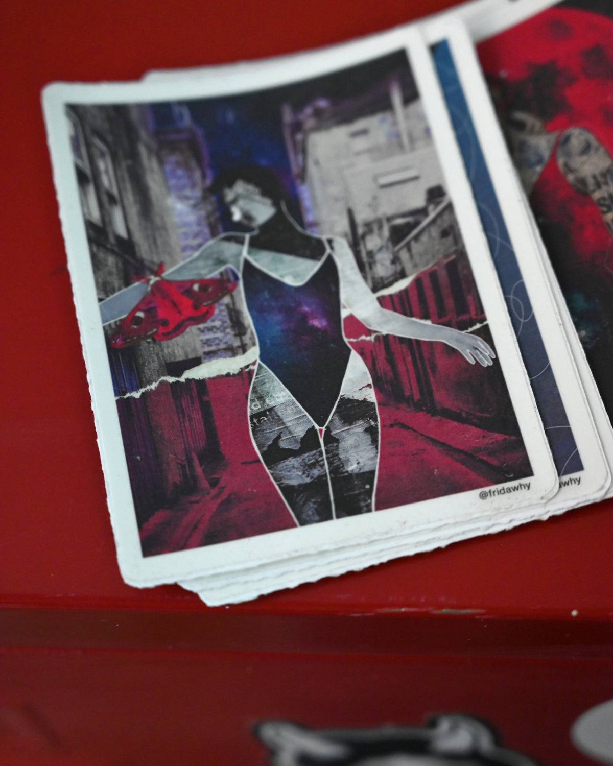 A close-up of a printed art card or sticker featuring a stylized collage portrait of a woman in a black bodysuit with red and blue abstract elements, resting on a red surface.