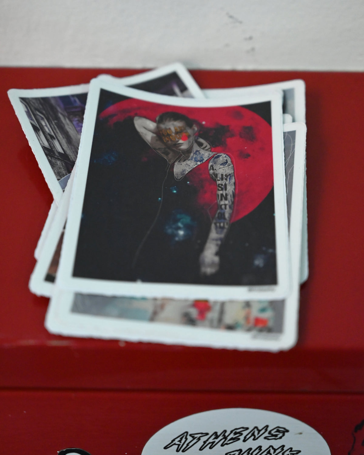 A stack of printed art cards or postcards with white borders, featuring a tattooed figure in front of a large red moon graphic, resting on a red surface.