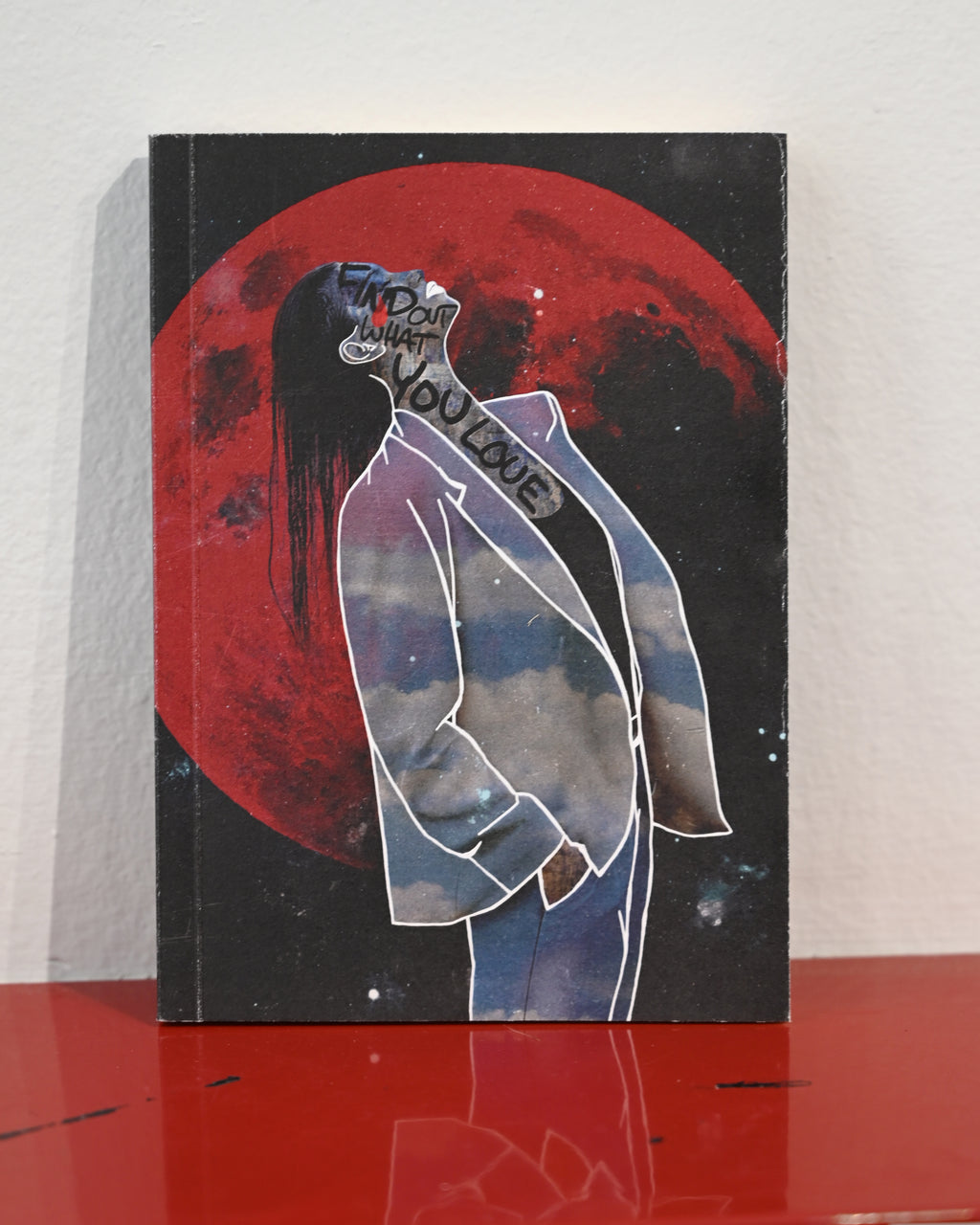 A square art print or book cover standing upright on a glossy red surface against a white wall. The artwork shows a stylized figure in a white suit outlined in white, with a large red moon or planet behind them on a black background.