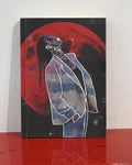 A square art print or book cover standing upright on a glossy red surface against a white wall. The artwork shows a stylized figure in a white suit outlined in white, with a large red moon or planet behind them on a black background.