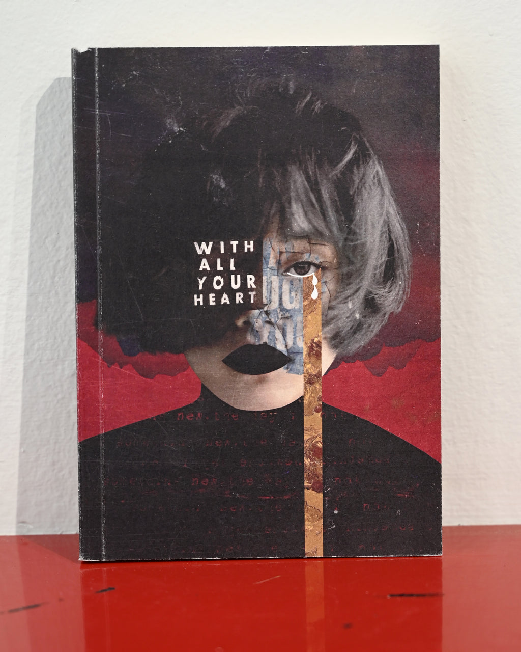A square art print or book cover standing upright on a red surface against a white wall. The artwork shows a stylized portrait with the text 'WITH ALL YOUR HEART' over a face obscured by dark hair, red abstract background shapes, and a vertical gold-toned textured stripe.