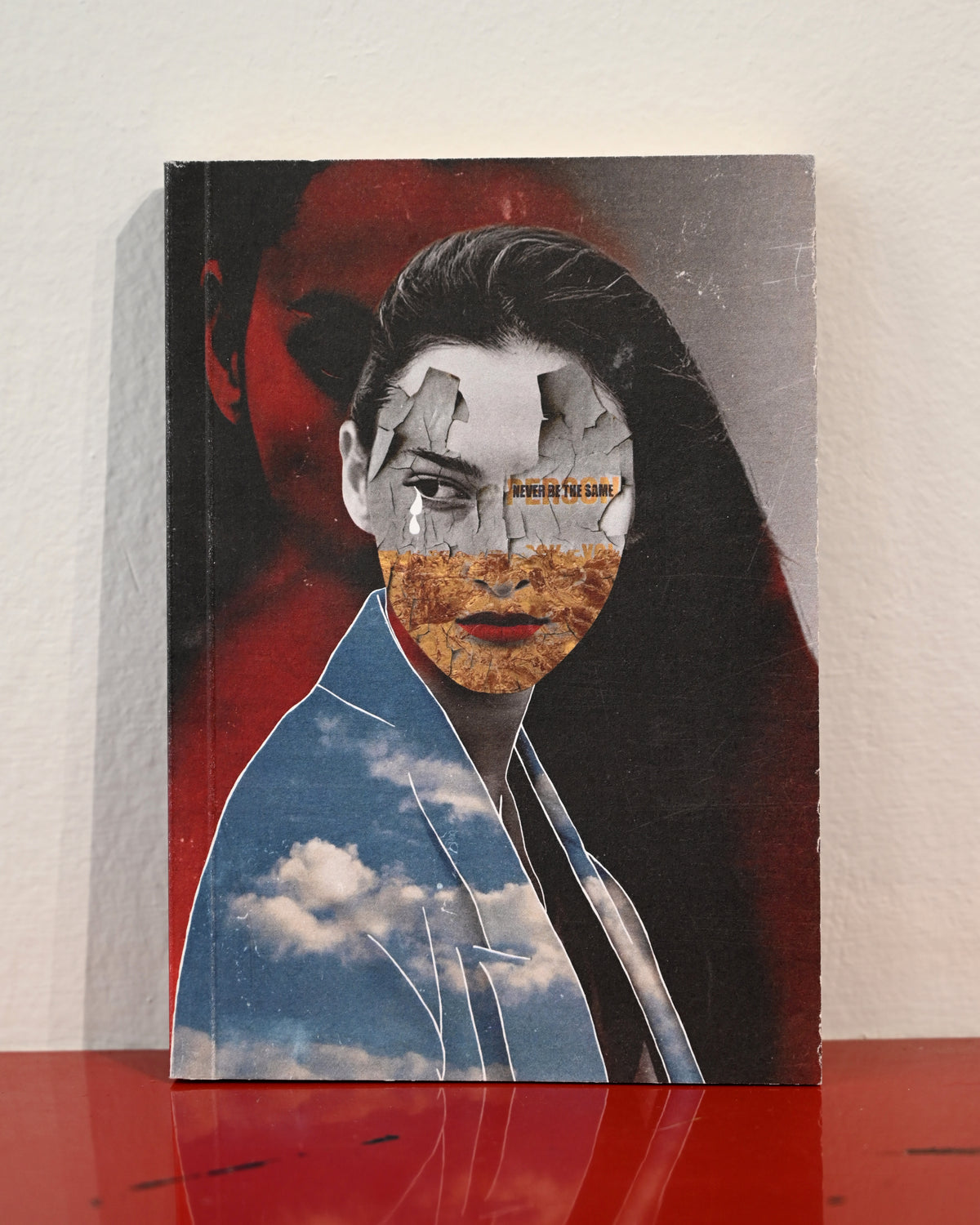 A square art print or book cover featuring a surreal portrait collage of a woman's face with cracked stone, orange-brown texture, and a blue sky with clouds over her shoulder, set against a red and black background. The item is photographed standing on a red reflective surface against a light wall.