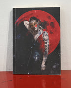 A hardcover notebook or journal standing upright on a glossy red surface against a white wall. The cover features a surreal illustration of a tattooed woman with her eyes obscured by a red square, set against a large red moon and starry space background.