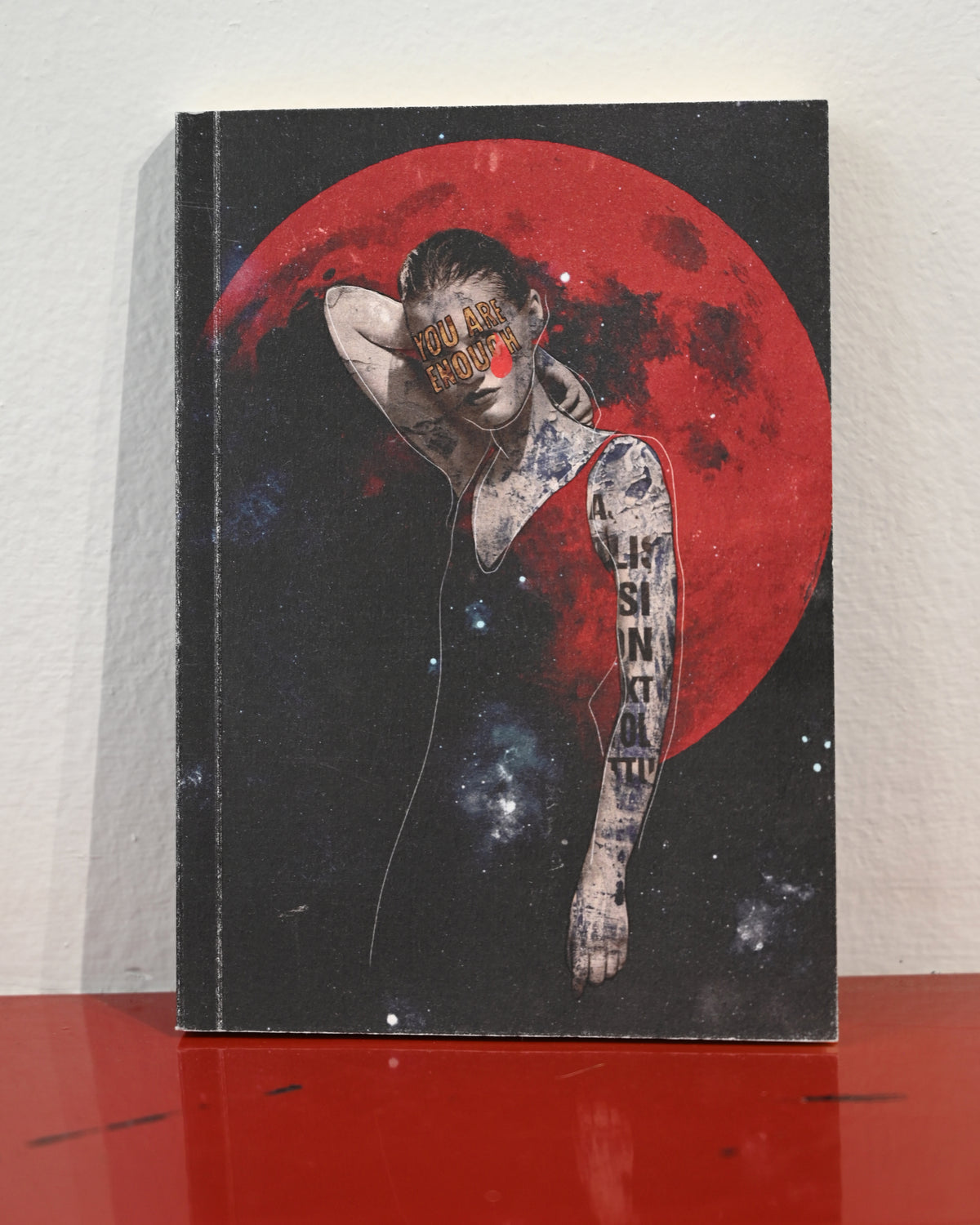 A hardcover notebook or journal standing upright on a glossy red surface against a white wall. The cover features a surreal illustration of a tattooed woman with her eyes obscured by a red square, set against a large red moon and starry space background.