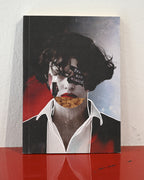 A hardcover art journal standing upright on a glossy red surface against a white wall. The cover features a surreal collage portrait of a woman with dark hair, a white collared shirt, red lips, and a pizza slice covering her mouth, with the text 'You are missed' on her hair.
